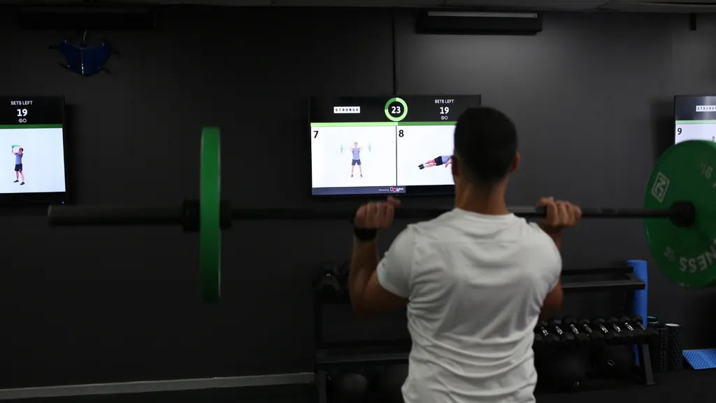 A gym member lifting a barbell in front of screens showing exercise demonstrations and a countdown
